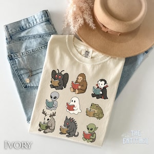 May include: An ivory t-shirt featuring a graphic print of nine cartoon creatures, including a mothman, bigfoot, vampire, alien, ghost, frog, wendigo, and goblin, each reading a book. The shirt is paired with light blue denim jeans and a tan hat.