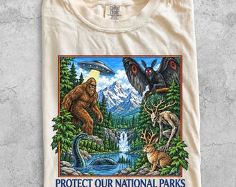 May include: An ivory t-shirt featuring a graphic print of a forest landscape with a UFO, Bigfoot, a moth-like creature, a deer-like entity, and a jackalope near a waterfall and lake. The text below the graphic reads PROTECT OUR NATIONAL PARKS.