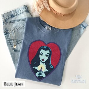 May include: A blue t-shirt featuring a graphic of a pale-skinned person with long black hair holding a lit candle inside a red heart shape. The shirt is paired with light blue denim jeans, a tan wide-brimmed hat, and a gold chain necklace.