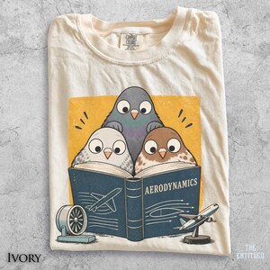 May include: An ivory t-shirt featuring a graphic of three birds reading a dark blue book titled AERODYNAMICS. The illustration includes a small wind tunnel fan and a model airplane. The shirt has a Comfort Colors brand label at the inner neckline.