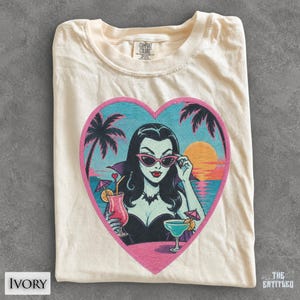 May include: An ivory t-shirt featuring a heart-shaped graphic of a pale-skinned person with black hair, wearing pink cat-eye sunglasses and a bat necklace. The scene depicts a tropical sunset with palm trees, cocktails, and umbrellas.
