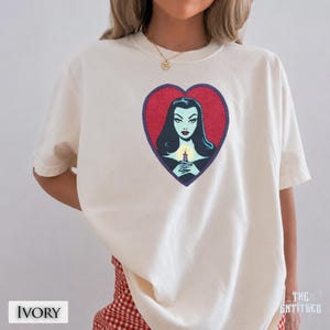 May include: An ivory short-sleeve t-shirt featuring a graphic print of a woman with long black hair and pale skin holding a lit candle. The illustration is framed within a red heart shape with a dark purple outline. The shirt has a crew neckline.