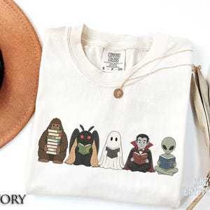 May include: An ivory t-shirt featuring a graphic of five characters reading books: a brown Bigfoot, a black Mothman, a white ghost, a vampire in a black cape, and a green alien. The shirt has a Comfort Colors label at the neck and a gold necklace.