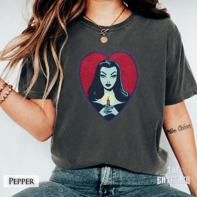 May include: A charcoal gray short-sleeve t-shirt featuring a graphic print of a pale-skinned person with long black hair inside a red heart shape. The person is holding a lit candle with a yellow flame. The shirt has a crew neckline and short sleeves.