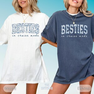 May include: Two oversized t-shirts, one white and one denim blue, each featuring the text BESTIES in a blue floral block font with the words in cruise mode written below in a cursive script. Both shirts have a crew neckline and short sleeves.