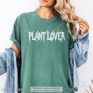 以下が含まれることがあります： 白のゴシック体で「PLANT LOVER」と書かれた、ライトグリーンのTシャツ。モデルはオーバーサイズフィットのためにXLサイズを着用しています。シャツは通常のSサイズです。