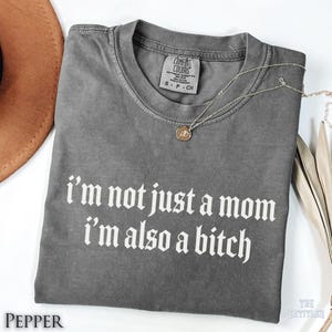 May include: A charcoal grey short-sleeve t-shirt with a crew neckline and a Comfort Colors brand label. The shirt features white text that reads, I'm not just a mom I'm also a bitch, printed in a bold, gothic-style font across the chest area.