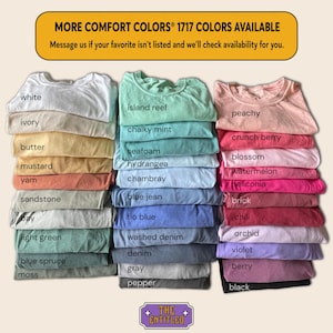 May include: A collection of folded Comfort Colors 1717 t-shirts arranged in three vertical stacks. The shirts display a wide variety of colors, each labeled with text including white, ivory, butter, mustard, yam, sandstone, bay, light green, blue spruce, moss, island reef, chalky mint, seafoam, hydrangea, chambray, blue jean, flo blue, washed denim, denim, gray, pepper, peachy, crunch berry, blossom, watermelon, heliconia, brick, chili, orchid, violet, berry, and black.