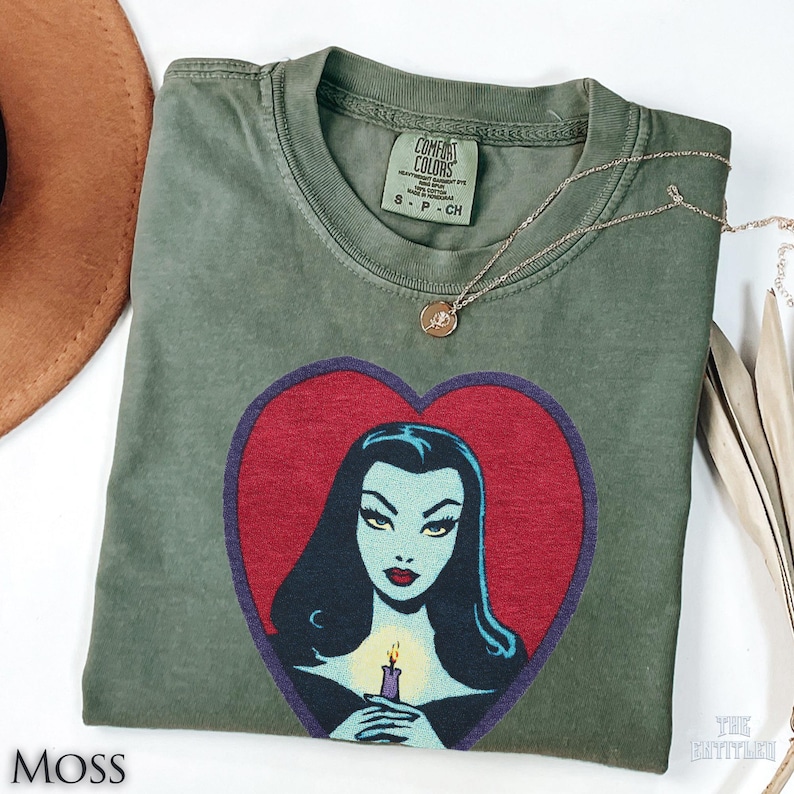 May include: A moss green short-sleeve t-shirt featuring a graphic print of a pale-skinned person with long black hair inside a red heart shape. The person holds a lit candle. The shirt has a Comfort Colors label at the neck and a crew neckline.