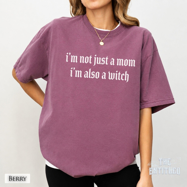 May include: A berry-coloured short-sleeve t-shirt featuring the white text "I'm not just a mom I'm also a witch" printed in a gothic-style font across the chest. The garment has a crew neckline and a relaxed fit, worn with a gold chain necklace.