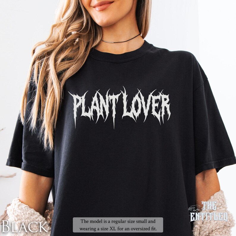 以下が含まれることがあります： 黒いTシャツに、白い「PLANT LOVER」の文字が尖ったフォントでプリントされています。「THE ENTITLED」のコレクションです。モデルはオーバーサイズフィットのためにXLサイズを着用しています。