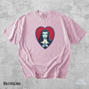 May include: A light pink short-sleeve t-shirt featuring a central graphic of a woman with dark hair holding a lit candle inside a red heart shape with a dark blue border. The shirt has a crew neckline and a standard fit with visible fabric texture.