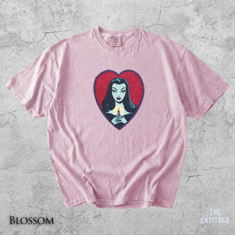 May include: A light pink short-sleeve t-shirt featuring a central graphic of a woman with dark hair holding a lit candle inside a red heart shape with a dark blue border. The shirt has a crew neckline and a standard fit with visible fabric texture.
