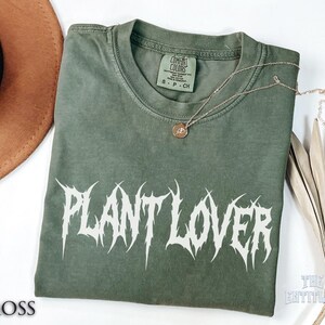 以下が含まれることがあります： モスグリーンのTシャツ。白いゴシックフォントで「PLANT LOVER」の文字がプリントされています。小さなペンダントが付いたゴールドのネックレスがTシャツの上にあります。Tシャツは白い表面に折りたたまれています。
