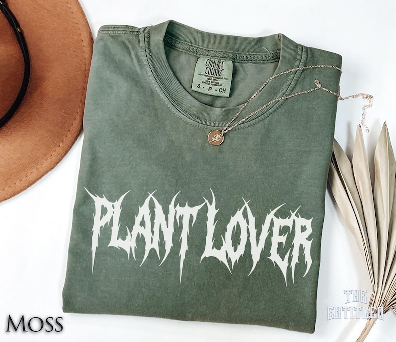 以下が含まれることがあります： モスグリーンのTシャツ。白いゴシックフォントで「PLANT LOVER」の文字がプリントされています。小さなペンダントが付いたゴールドのネックレスがTシャツの上にあります。Tシャツは白い表面に折りたたまれています。