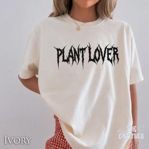 以下が含まれることがあります： アイボリー色のTシャツ。テキスト「PLANT LOVER」は、とげのあるゴシックフォントで書かれています。 ゆったりとしたフィット感で、半袖です。「THE ENTITLED」の文字が右下に印刷されています。
