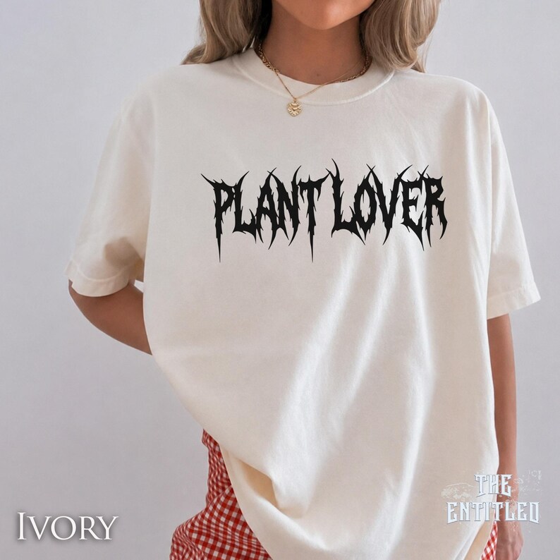 以下が含まれることがあります： アイボリー色のTシャツ。テキスト「PLANT LOVER」は、とげのあるゴシックフォントで書かれています。 ゆったりとしたフィット感で、半袖です。「THE ENTITLED」の文字が右下に印刷されています。