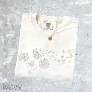 May include: An ivory t-shirt featuring a graphic of three dandelion-like flowers composed of medical instruments, including stethoscopes, syringes, and pills. A gold-toned necklace with a circular pendant rests on the neckline of the shirt.