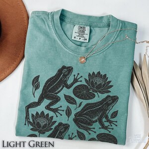 May include: A light green short-sleeve t-shirt featuring a black ink-style graphic print of two frogs, lily pads, and lotus flowers. The shirt has a crew neckline and a Comfort Colors brand label on the inside collar, size small.