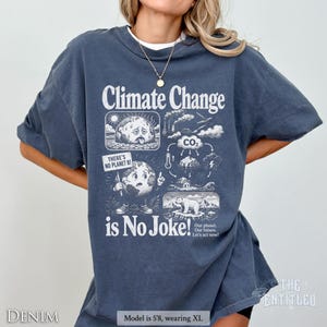 May include: A dark blue t-shirt featuring a white graphic print with the text Climate Change is No Joke! The design includes illustrations of a sad Earth, a CO2 cycle diagram, a polar bear on ice, and a sign that reads There's No Planet B.