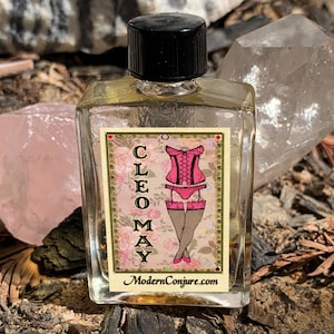May include: A small clear glass bottle with a black cap, containing a light yellow liquid. The bottle has a label with the text "CLEOMAY" and a pink illustration of a woman wearing a corset and stockings. The label also includes the text "ModernConjure.com".