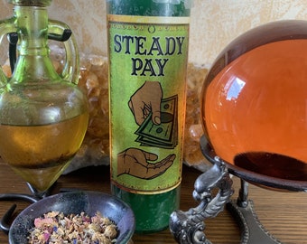 May include: Green candle with a label that reads "Steady Pay" and depicts a hand receiving money. The candle is on a wooden surface with a small bowl of dried herbs and a decorative stand with a glass orb.