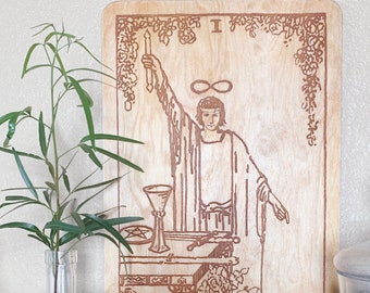 May include: A wooden tarot card with a brown background and a brown illustration of The Magician. The card is surrounded by a decorative border of flowers and vines. The text "THE MAGICIAN." is printed at the bottom of the card.