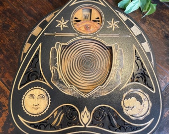Könnte beinhalten: Ein schwarzes, dreieckiges Holzbrett mit einem dekorativen Design, das eine Spirale, ein Auge und zwei Hände zeigt. Das Brett hat eine goldfarbene Oberfläche und ist mit komplizierten Details verziert.
