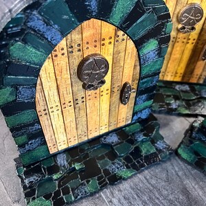 May include: A miniature wooden door with a stone archway, painted in shades of green and black. The door has a metal handle and a decorative metal circle with a design in the center. The door is set on a base of green and black mosaic tiles.