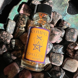 May include: A small, clear glass bottle with a black cap filled with a golden liquid. The bottle has a yellow label with the words "COMPEL SPELL OIL BY MODERN WITCH" and a pentagram symbol. The bottle is sitting on a bed of pink and black stones.