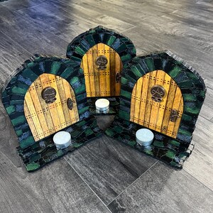 May include: Three wooden door-shaped candle holders with a green and black mosaic design. Each holder has a small, round, white candle inside.