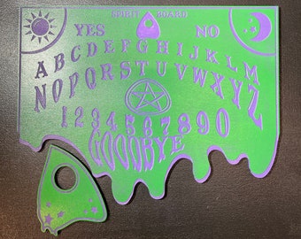 Könnte beinhalten: Grünes und violettes Ouija-Brett mit einem Planchette. Das Brett hat Buchstaben, Zahlen und die Wörter "YES", "NO" und "GOODBYE".