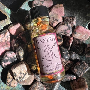 May include: A small glass bottle with a black cap filled with a golden liquid. The label reads "Banish Spell Oil" with a hand with lightning bolts around it. The bottle is sitting on a bed of pink and black stones.