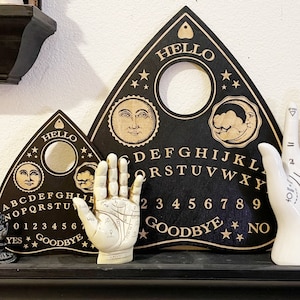 May include: Two black wooden Ouija boards with gold lettering and symbols. The larger board has the words "HELLO" and "GOODBYE" at the top and bottom. The smaller board has the words "HELLO" and "GOODBYE" at the top and bottom. A white ceramic hand with lines and symbols is in front of the boards.
