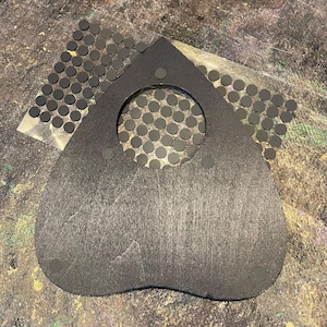 May include: A heart-shaped black planchette with a circular cutout in the center. The planchette features three small circular adhesive pads attached to its surface. A sheet of numerous small, round, black adhesive dots is positioned behind it.