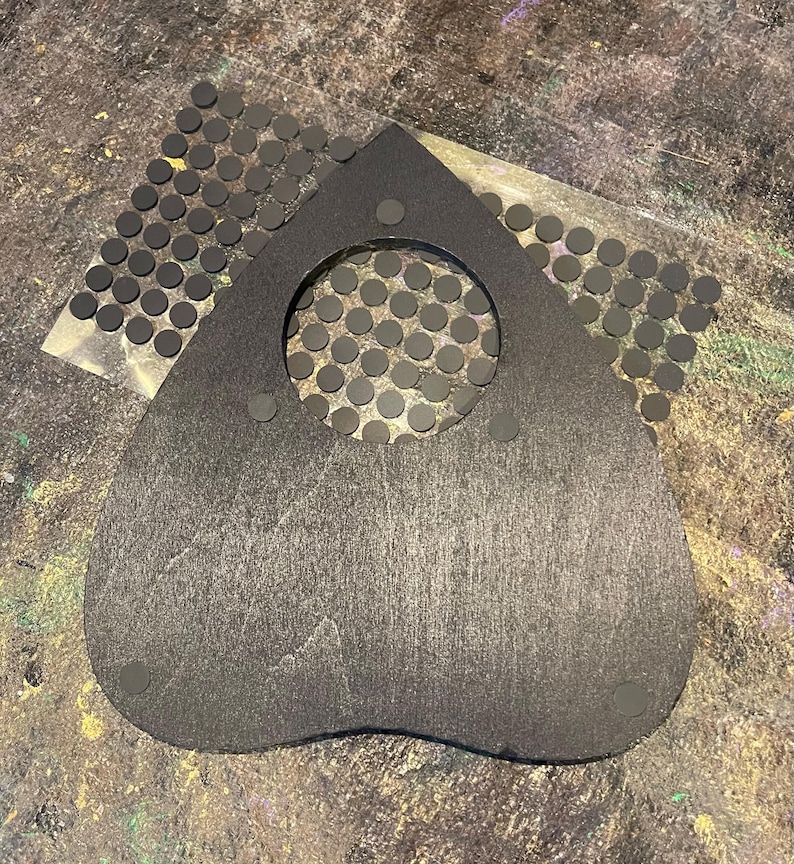 May include: A heart-shaped black planchette with a circular cutout in the center. The planchette features three small circular adhesive pads attached to its surface. A sheet of numerous small, round, black adhesive dots is positioned behind it.