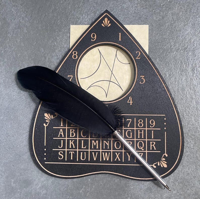 May include: A black heart-shaped spirit board with copper-colored lettering featuring the alphabet and numbers one through nine. A circular cutout at the top reveals a beige insert with black line art. A black feather quill pen rests on top.