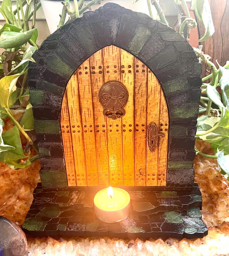 May include: A miniature wooden door with a stone archway and a small candle in front of it. The door is painted to look like wood and has a metal handle. The archway is painted to look like stone and has a green and black color scheme.