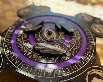 Könnte beinhalten: Ein Holz-Divinationsbrett mit einem violetten Zentrum und einem drehbaren Zifferblatt. Das Zifferblatt hat einen schwarzen Hintergrund mit goldenen Buchstaben und Symbolen. Das Brett hat drei Holzhandflächen mit Augen, die jeweils auf ein anderes Symbol auf dem Zifferblatt zeigen. Der Text auf dem Zifferblatt lautet "Otho".