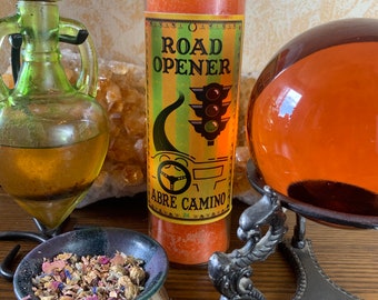 May include: A red and orange candle with a yellow label that reads "Road Opener" and "Abre Camino". The label also features a traffic light and a steering wheel. The candle is on a wooden surface with a small bowl of dried flowers and a glass orb on a black metal stand.