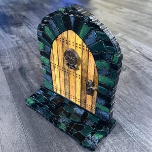 May include: A miniature wooden door with a green and blue mosaic frame and a yellow wooden door with a metal handle and a decorative metal piece. The door is on a base made of green and blue mosaic tiles.