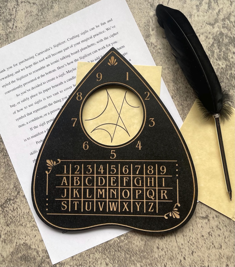 May include: A black wooden planchette-shaped tool featuring a circular cutout, engraved numbers one through nine, and a grid containing the alphabet from A to Z. The surface displays gold-colored etched lines and text, accompanied by a black feather pen.