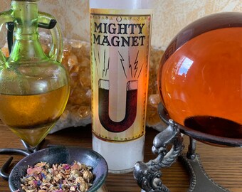 May include: White candle with a label that reads "Mighty Magnet" and a graphic of a horseshoe magnet with lightning bolts. The candle is on a wooden surface with a small bowl of dried herbs and a glass orb on a stand.