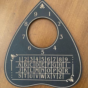 May include: A black teardrop-shaped planchette featuring a circular cutout at the top. The surface is engraved with the numbers 1 through 9 arranged in a circle around the cutout, and a grid below containing the numbers 1 through 9 and letters A through Z.