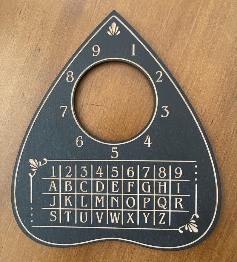 May include: A black teardrop-shaped planchette featuring a circular cutout at the top. The surface is engraved with the numbers 1 through 9 arranged in a circle around the cutout, and a grid below containing the numbers 1 through 9 and letters A through Z.