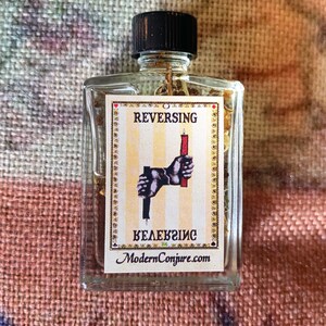 May include: A clear glass bottle with a black cap, containing a yellow liquid. The label on the bottle reads "REVERSING" with a graphic of two hands holding candles. The text "ModernConjure.com" is also on the label.