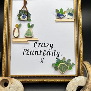 May include: Framed artwork in a gold-coloured ornate frame. The artwork has a white background with small, colourful glass pieces arranged to resemble plants. The text "Crazy Plant Lady x" is written in black script.