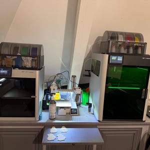 May include: Two 3D printers are displayed on a gray cabinet. The printer on the left is silver with a black door and a screen. The printer on the right is white with a black door and a green light inside. A smaller printer is in the middle.
