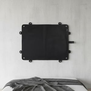 Könnte beinhalten: Ein rechteckiges, schwarzes Wandobjekt mit D-Ringen zum Aufhängen. Es ist an einer weißen Wand über einem Bett mit einer grauen Decke angebracht. Das Objekt hat ein minimalistisches Design.