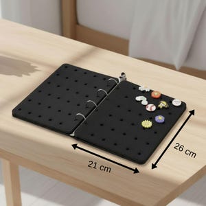 Puede incluir: Un archivador negro de fieltro con numerosos agujeros pequeños, abierto sobre una superficie de madera clara. El archivador mide 21 cm de ancho y 26 cm de largo. Varios alfileres decorativos están adheridos al lado derecho.